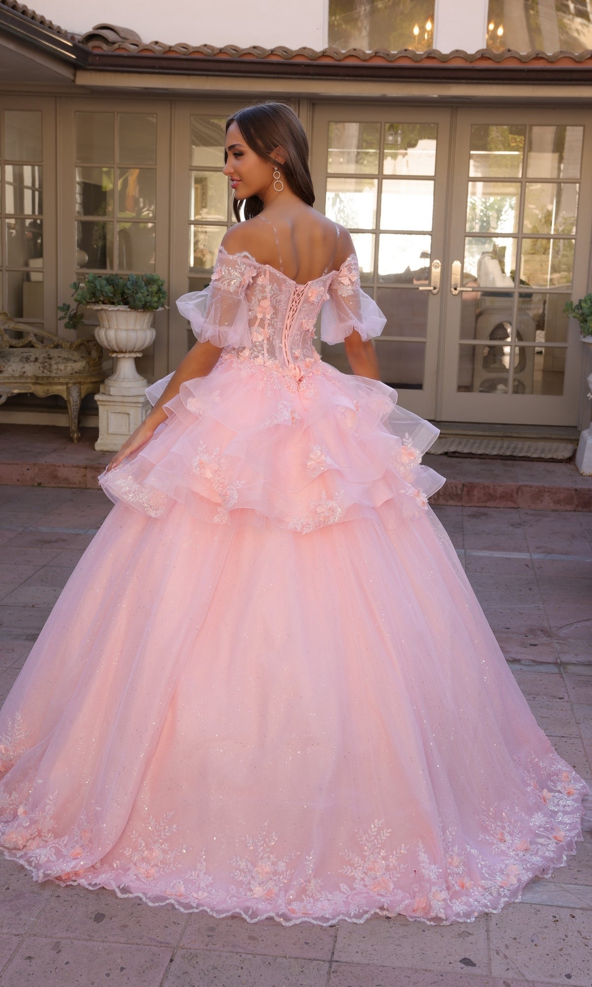 A young woman stands outdoors, facing away, wearing the Nox Anabel Removable-Skirt Formal Ball Gown H1360—a light pink off-the-shoulder dress with layered tulle and floral embroidery.